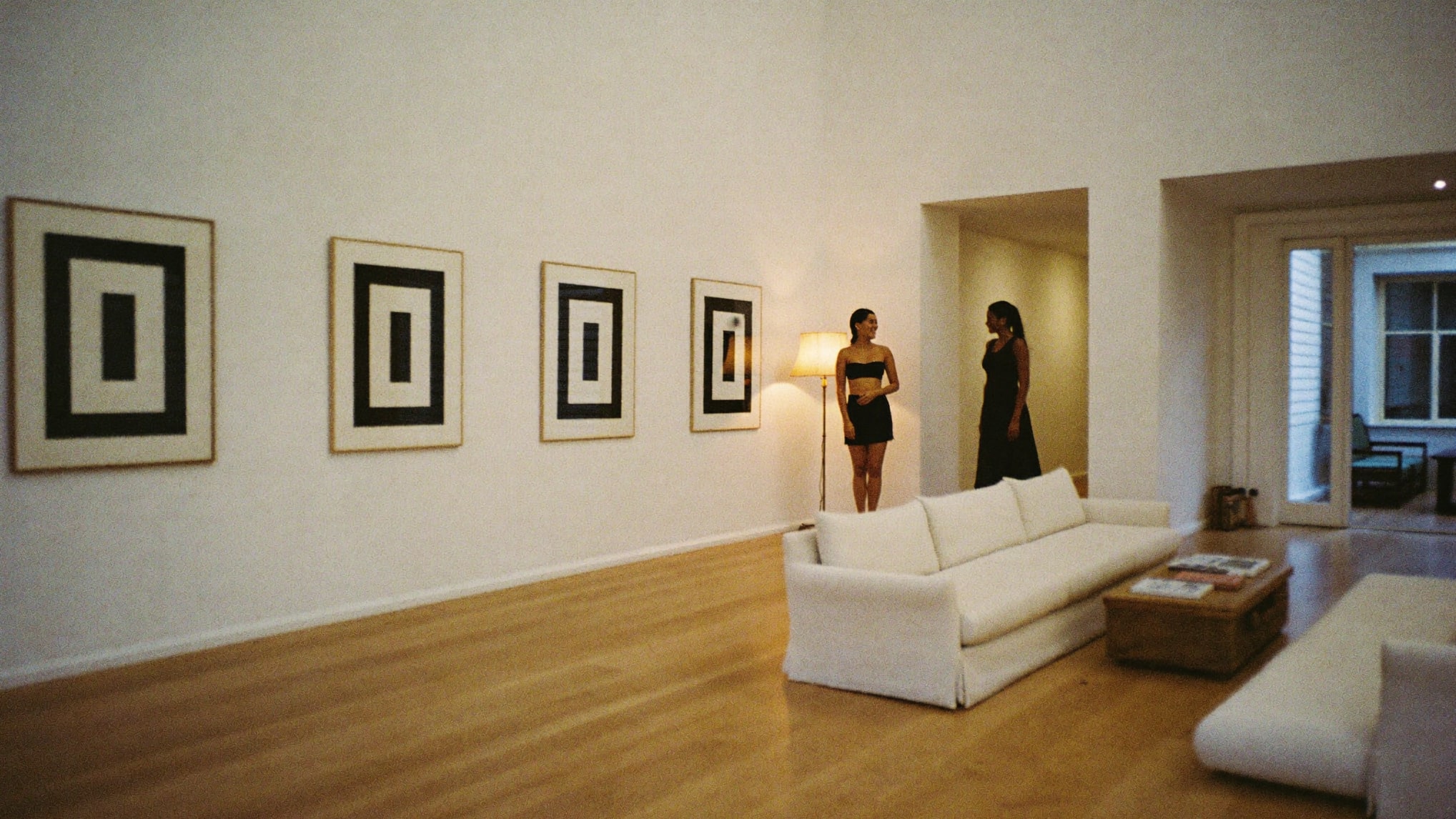 Two women talking at dusk in a room hung with three framed works.