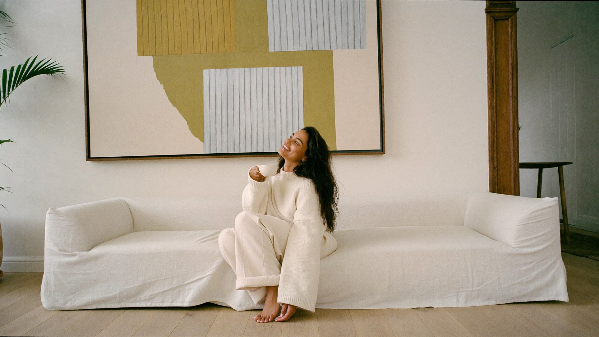 A woman curled into a linen sofa with tea, looking up at a large framed painting.
