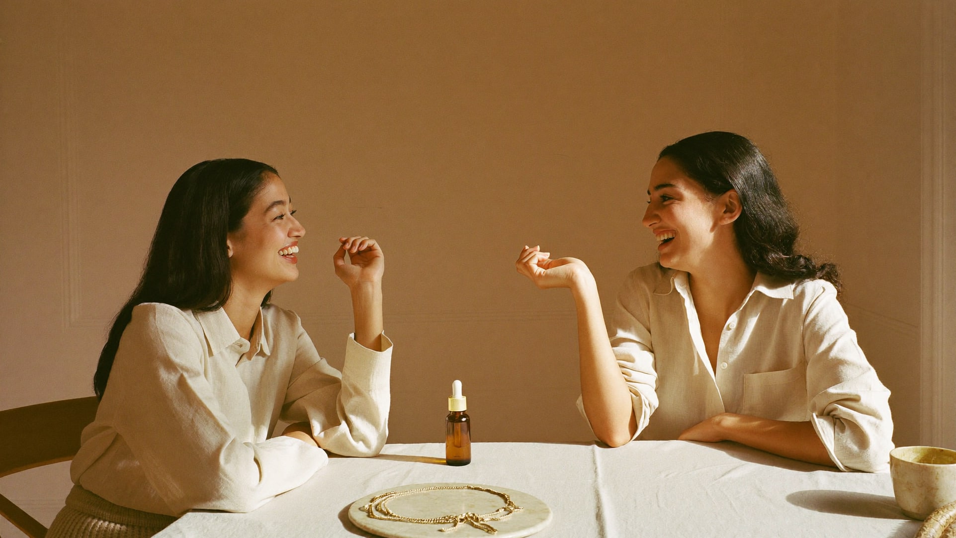 Two women laughing over a table set with a glass bottle and a gold chain.